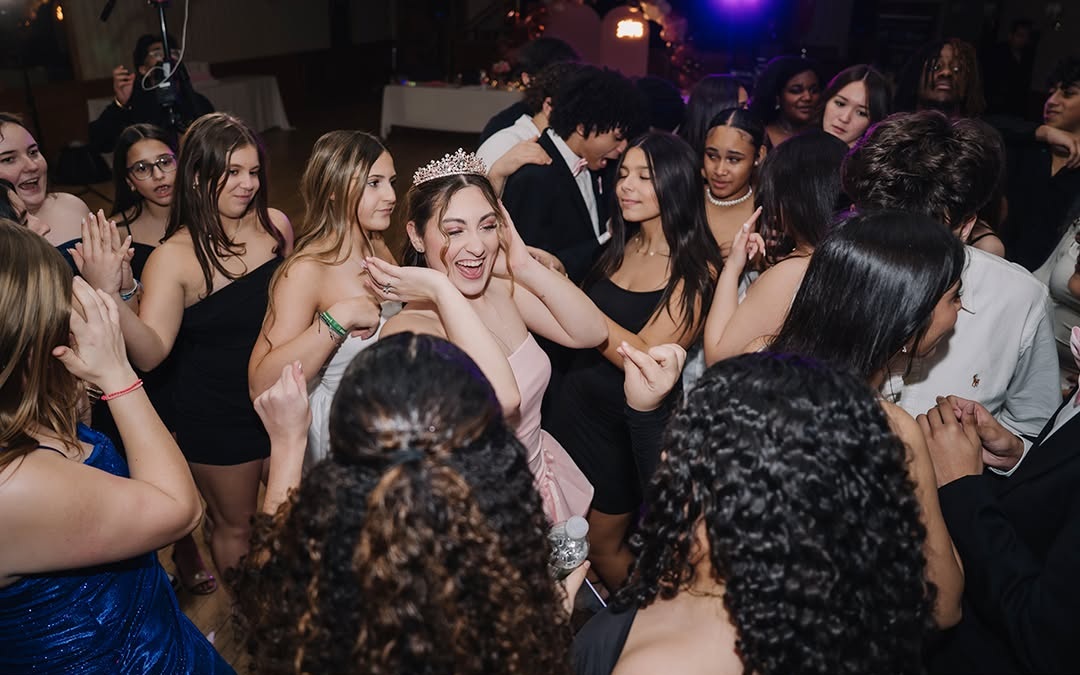 Smiling Sweet 16 birthday girl wearing a tiara and pink dress dances with friends at a lively party, captured in a candid celebration photograph.