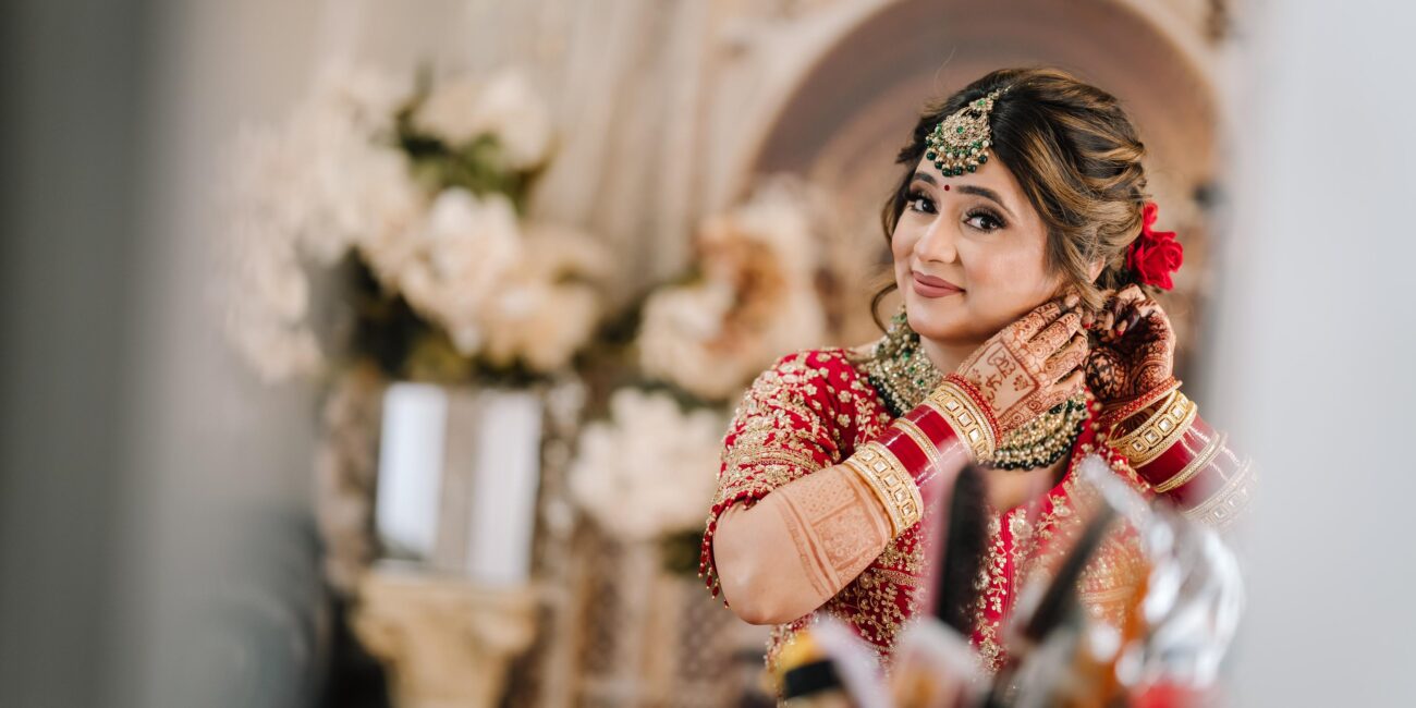 Bride adjusting earrings in bridal suite, detailed red and gold bridal attire and henna — captured by Citi Events Productions.
